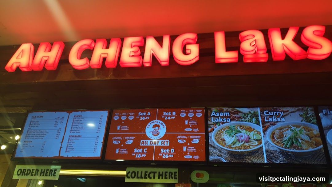A brightly lit storefront featuring a large red neon sign reading 'AH CHENG LAKSA'. Below the sign, there are illuminated menu boards displaying various food items and prices. The left board lists all-carat dishes, snacks, and beverages. The center board highlights set meals like Set A, B, C, D, and an All Day Set. The right board showcases images of Asam Laksa and Curry Laksa with their respective names.