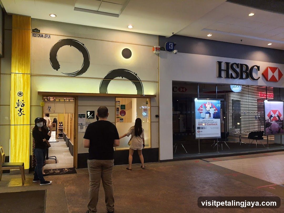 The image shows the exterior entrance of a restaurant with Japanese-style decor, including circular wall art and vertical wooden panels. A couple stands near the entrance, while another person is visible inside. Adjacent to the restaurant is an HSBC bank branch with promotional posters in the window.