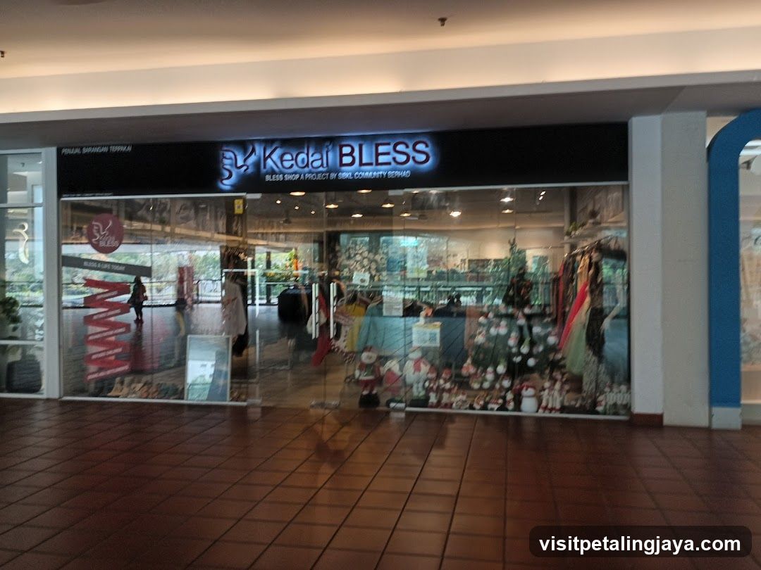A shop front with a black signboard displaying 'Kedai BLESS' in illuminated white letters. The storefront features large glass windows and doors, revealing an interior display of merchandise including clothing racks, decorative items, and small figurines. The floor is tiled, and the entrance appears to be accessible from a mall corridor.