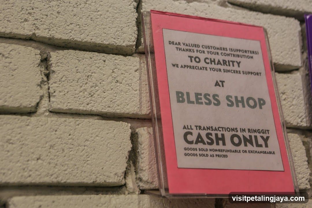 A pink-framed sign mounted on a textured beige brick wall outside a shop. The sign reads: 'Dear valued customers (supporters) thanks for your contribution to charity we appreciate your sincere support at BLESS SHOP all transactions in ringgit cash only goods sold non-refundable or exchangeable goods sold as priced'. The text is printed in black and grey on white paper, enclosed in clear plastic.
