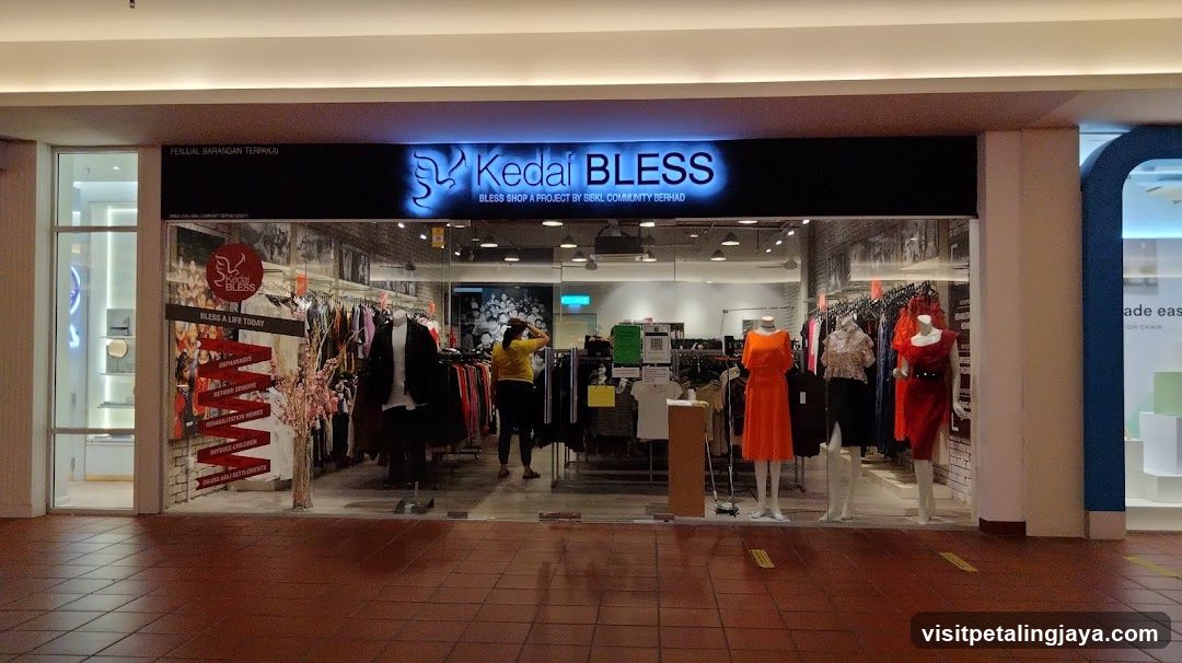 A brightly lit retail storefront with a glowing blue neon sign reading 'Kedai BLESS' and the tagline 'Bless Shop A Project by Serik Community Berhad'. The shop features large glass windows displaying mannequins in colorful clothing, including orange dresses and patterned tops. Inside, racks of clothes are visible along with promotional posters on the left wall. The floor is tiled, and the entrance is flanked by white pillars.
