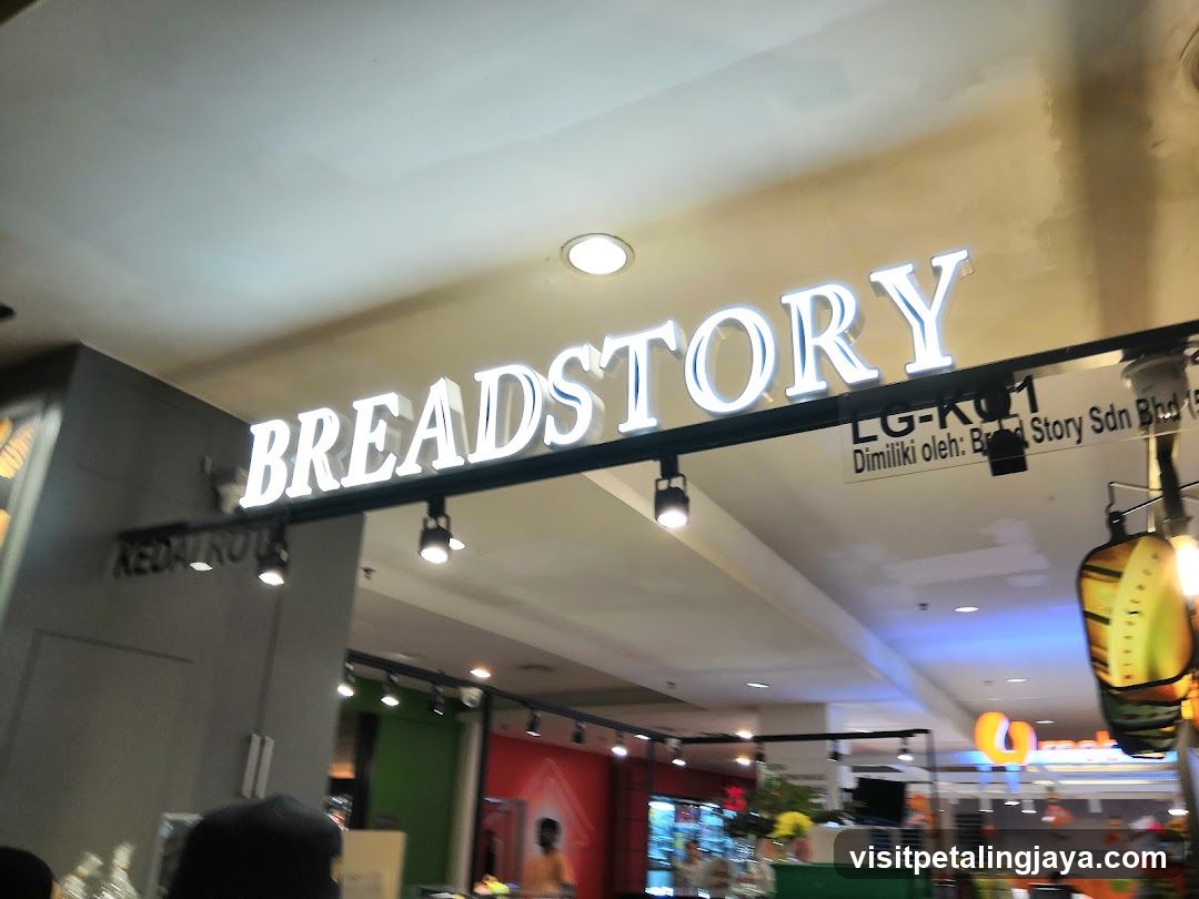 The image shows the storefront of a Breadstory bakery, with its illuminated sign prominently displayed above the entrance. The store is located in a shopping mall or commercial area, as indicated by the surrounding architecture and lighting. There are no visible menus or price lists in the image.