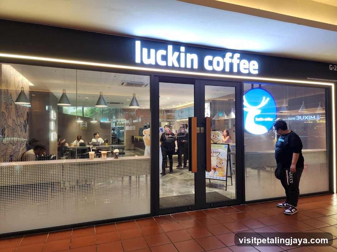A Luckin Coffee shop with illuminated signage above the entrance, glass doors and windows showing interior staff and customers, a promotional standee near the door, and a customer standing outside.
