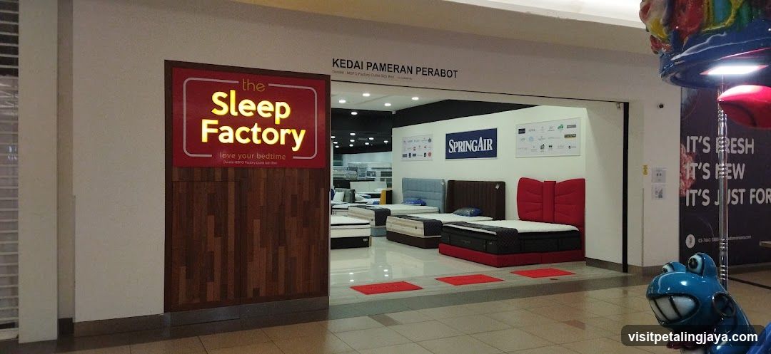 A photo of the physical exterior entrance to The Sleep Factory, a furniture store specializing in mattresses and beds. A large illuminated sign with yellow lettering on a red background is mounted on the building facade. Inside, various bed models are displayed against white walls, including brands like SpringAir. Red floor mats lead into the showroom.