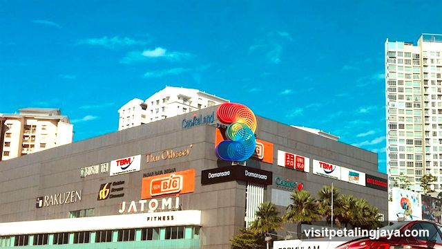 A modern multi-story commercial building features numerous visible brand logos and signage on its exterior facade, including Rakuzen, Jatomi Fitness, Danamart, TBM, and others. The structure is set against a clear blue sky with surrounding high-rise buildings in the background.